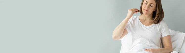 Woman in a white t-shirt sitting in bed with fresh sheets and applying a Saje roll-on to her neck