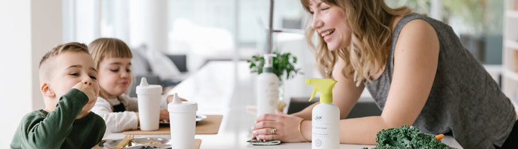 A mother watching her two young children eat on a white kitchen island, with Saje home cleaning bottles placed next to her