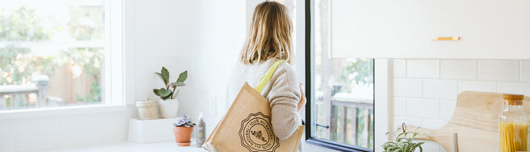 Woman in a bright white kitchen carrying a Saje jute tote bag beside a Saje room spray resting on the counter