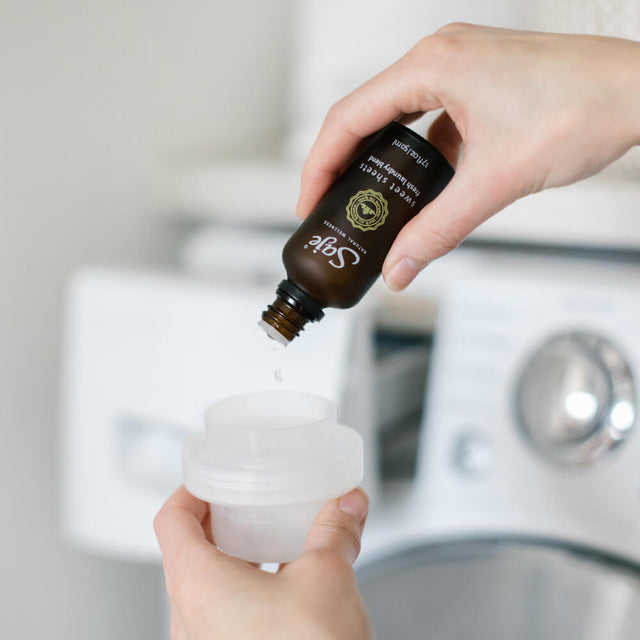 A person adding drops of Sweet Sheets laundry blend to a cap of laundry detergent